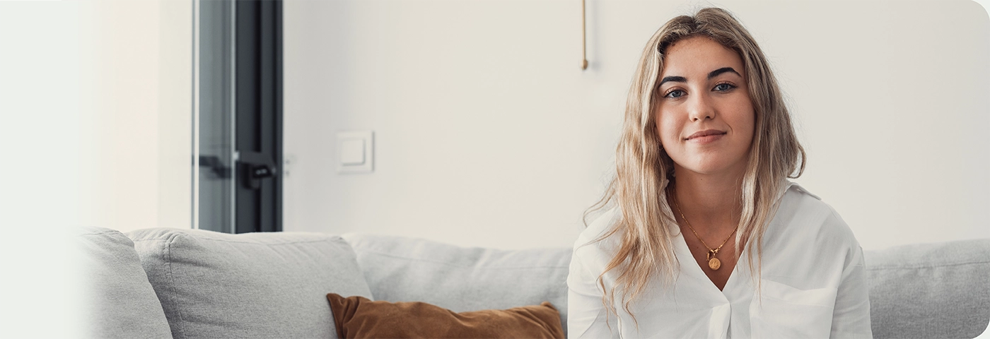 Woman sitting on couch in light apartment with small smile on her face