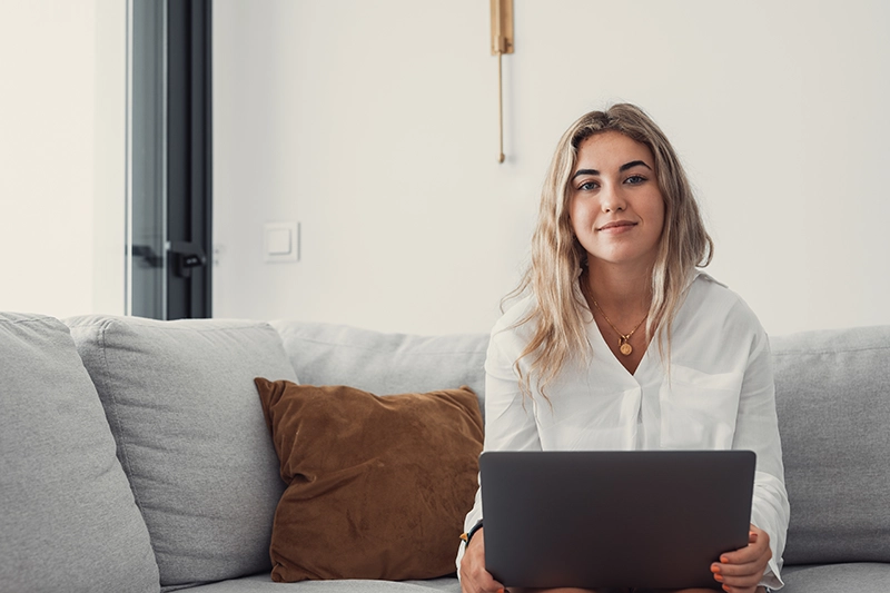 Woman sitting on couch with laptop in light apartment with small smile on her face